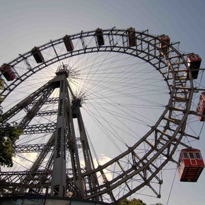 May include: A large, metal Ferris wheel with red passenger cars. The wheel is against a blue sky with white clouds.