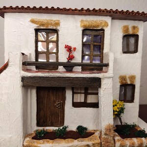 May include: A white miniature house with a brown roof, a wooden door, and windows. The house has a small balcony with a potted plant. There are small bushes and a stone path in front of the house.