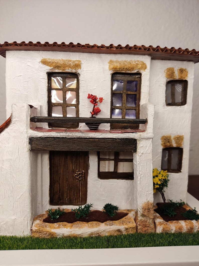 May include: A white miniature house with a brown roof, a wooden door, and windows. The house has a small balcony with a potted plant. There are small bushes and a stone path in front of the house.