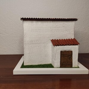 May include: A white miniature house with a red tile roof and a brown wooden door. The house is sitting on a white base with green grass.
