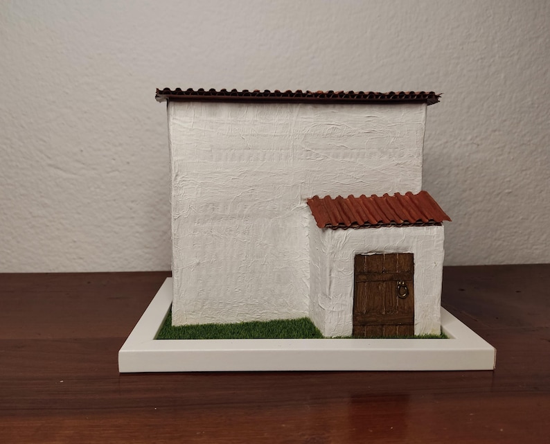 May include: A white miniature house with a red tile roof and a brown wooden door. The house is sitting on a white base with green grass.