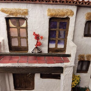 May include: A miniature white building with a red tile roof, two windows, a brown door, and a small red plant on a balcony. The building is made of paper and has a textured surface.