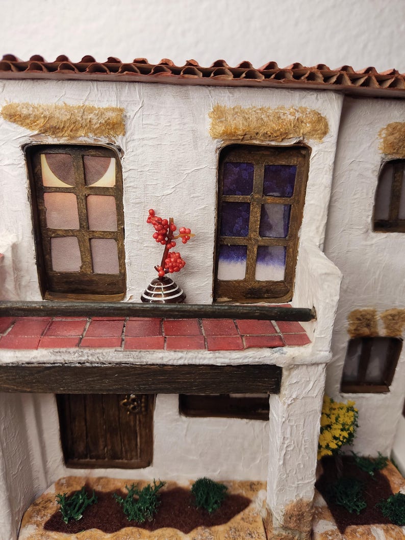 May include: A miniature white building with a red tile roof, two windows, a brown door, and a small red plant on a balcony. The building is made of paper and has a textured surface.