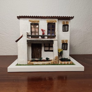 May include: A white miniature house with a red tile roof, brown wooden door, and windows. The house has a small balcony with a potted plant and a small bush in front.