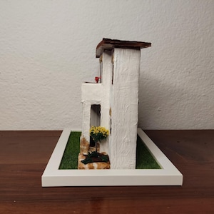 May include: A white miniature model of a two-story house with a brown roof, a small balcony, and a green lawn. The house has a white picket fence and a small flower pot with yellow flowers.