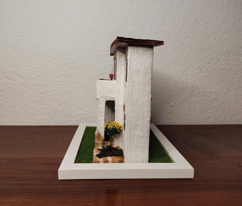 May include: A white miniature model of a two-story house with a brown roof, a small balcony, and a green lawn. The house has a white picket fence and a small flower pot with yellow flowers.