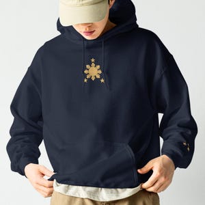 May include: A navy blue hooded sweatshirt featuring a gold embroidered sun and star emblem on the chest, paired with a beige baseball cap and khaki cargo trousers. The hoodie includes a front kangaroo pocket and long sleeves with ribbed cuffs.