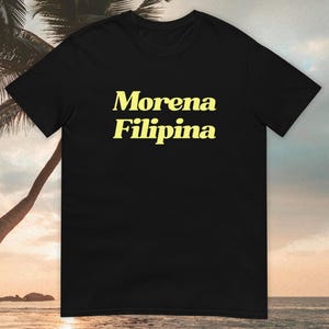 May include: Black t-shirt featuring the words "Morena Filipina" in a yellow, bold font. The image shows the shirt against a beach scene with palm trees and a sunset.