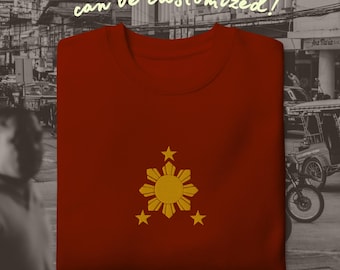 May include: A dark red crewneck sweatshirt featuring an embroidered gold sun and three stars emblem centered on the chest. The text "can be customized!" is printed in a light yellow script font above the garment against a blurred background.