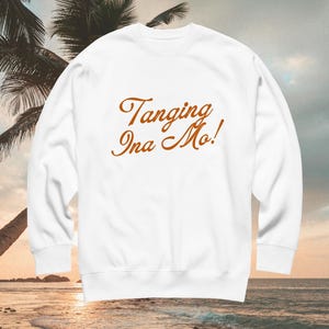 May include: A white long-sleeve sweatshirt featuring the text "Tanging Ina Mo!" printed in a brown, cursive-style font across the center of the chest. The garment has a crew neckline, long sleeves with ribbed cuffs, and a ribbed hemline.