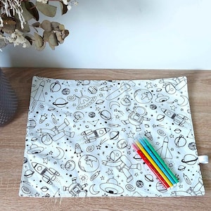 Peut inclure: Un set de table blanc avec un motif spatial noir et blanc. Le set de table représente des fusées, des planètes, des étoiles, des astronautes et des soucoupes volantes. Il y a cinq marqueurs colorés sur le dessus du set de table.