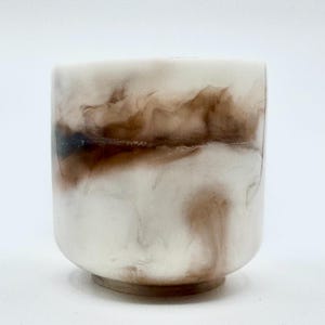 Pode incluir: Uma pequena chávena cilíndrica com base arredondada. A chávena apresenta um design marmoreado com redemoinhos de branco e castanho, criando um padrão orgânico único. A chávena é feita de um material brilhante.