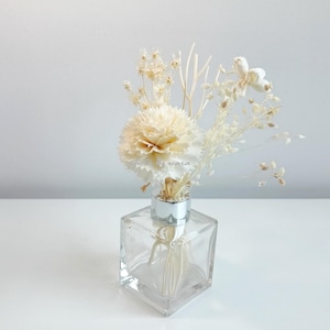 May include: A clear glass diffuser bottle with a silver cap and a bouquet of dried white flowers. The flowers include carnations, sprigs of dried grasses, and small white flowers.