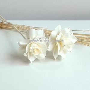 May include: Two white wooden flowers with a bundle of natural brown sticks. The flowers are arranged on a white surface.