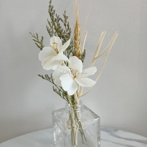 May include: A clear glass diffuser bottle with dried white flowers, wheat stalks, and twigs. The bottle is square shaped and sits on a white marble surface.