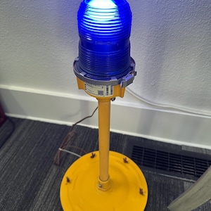 May include: A bright blue, ribbed, cylindrical beacon light mounted on a yellow metal pole with a circular yellow base. The beacon features a metallic silver collar and a white label with a QR code and text, connected by a thin brown electrical cord.