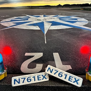 May include: Two white rectangular signs with blue lettering reading "N761EX" on a black asphalt surface. A blue and white compass rose is painted on the surface. Two blue and white flashing lights are on either side of the signs.