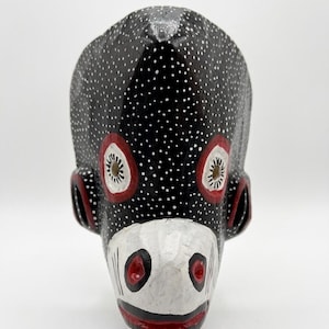 May include: A hand-carved wooden mask featuring a black surface covered in small white dots. The mask has a white snout area with black and red details, prominent red-rimmed eye sockets with circular cutouts, and red-accented ear-like side features.