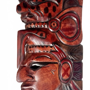 May include: A carved wooden mask with a layered design. The mask features a jaguar at the top with a spotted pattern, a central face with a prominent nose, and intricate details in shades of red and brown.