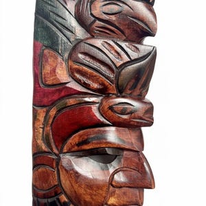 May include: A carved wooden totem pole with a dark brown finish. The pole features detailed carvings of birds and human faces, with accents of red and green. The artwork is a vertical design, showcasing traditional Native American artistry.