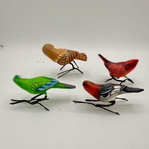 May include: Four small bird figurines with painted details, each featuring thin black wire legs. The birds are colored in shades of brown, bright red, vibrant green with blue accents, and a combination of red, white, and black on their bodies.
