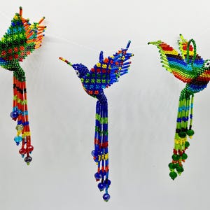 May include: Three colorful beaded hummingbird ornaments with long beaded tails. The hummingbirds are made of small glass beads in shades of green, blue, red, yellow, and orange. The tails are made of beads in the same colors.