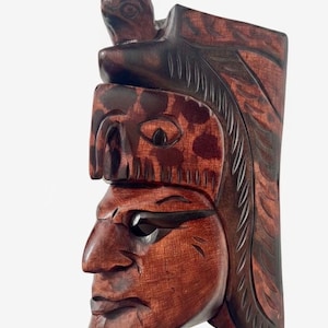 May include: A carved wooden mask depicting a Native American figure in profile. The mask is primarily brown with darker accents, featuring a detailed face, headdress, and a small bird carving at the top. The carving has a rustic, handcrafted appearance.
