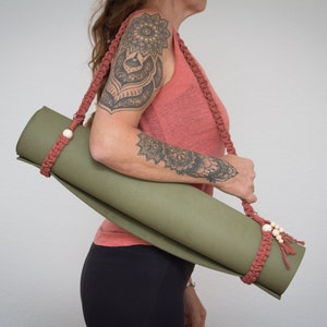 Könnte beinhalten: Eine Person trägt eine grüne Yogamatte mit einem roten gehäkelten Gurt und Holzperlen. Der Gurt ist verstellbar und hat einen Quasten am Ende.