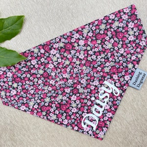 May include: A black bandana with a pink and white floral pattern. The bandana has the name "Daisy" embroidered on it in white. The bandana has a small white tag that says "Dashing Dogwear".