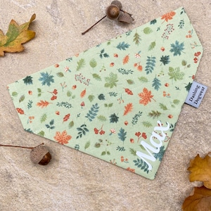 May include: A pale green dog bandana featuring a pattern of autumn leaves in shades of orange, green, and blue. The bandana is personalised with the name "Max" and a "Dashing Dogwear" label.