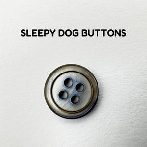 May include: A circular button with a metallic finish and four holes. The button has a distressed appearance and is set against a white backdrop. The text "SLEEPY DOG BUTTONS" is above it.