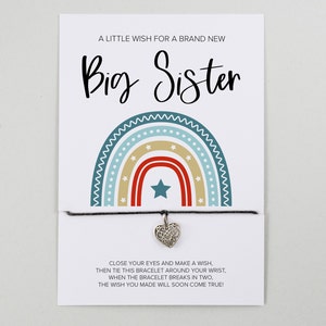 May include: A white card with a rainbow design and the text "A Little Wish for a Brand New Big Sister". A black string bracelet with a silver heart charm is attached to the card. The card includes the text "Close your eyes and make a wish, then tie this bracelet around your wrist. When the bracelet breaks in two, the wish you made will soon come true!"