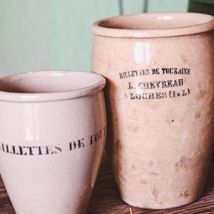 May include: Two antique beige ceramic jars with black text, one cylindrical and one tapered, sit on a wooden surface. A green glass bottle with flowers is in the background. The jars have a rustic, vintage aesthetic.