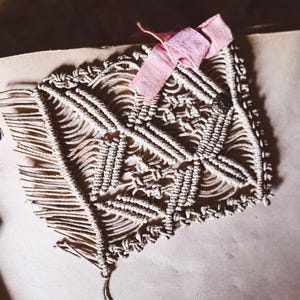May include: Handmade macrame clutch in a natural off-white color, featuring a geometric pattern. A delicate pink ribbon bow adorns the top. The clutch is a unique accessory for carrying essentials.