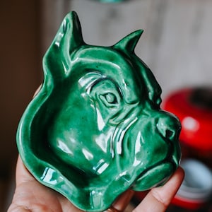 Könnte beinhalten: Eine glänzende, smaragdgrüne Keramikschale in Form eines Hundekopfes. Die Schale weist detaillierte Gesichtszüge auf, darunter Augen, Nase und Ohren. Die hundeförmige Schale ist ein dekorativer Gegenstand.
