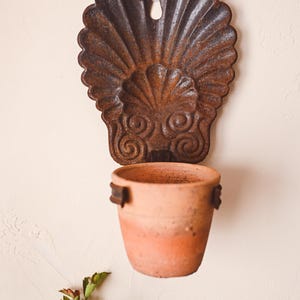 Pode incluir: Um vaso de parede de metal enferrujado em forma de concha de vieira com um vaso de terracota pendurado.
