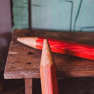 Könnte beinhalten: Zwei übergroße Holzstifte, einer davon im Fokus, liegen auf einer rustikalen Holzoberfläche. Die Stifte haben eine Spitze aus Naturholz und einen leuchtend roten Körper. Der Hintergrund zeigt eine verwitterte, türkis farbene Wand.