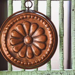 Peut inclure: Une décoration murale ronde en cuivre avec un motif floral en relief. La décoration est fixée à une surface en bois vert à lattes avec de la peinture écaillée.