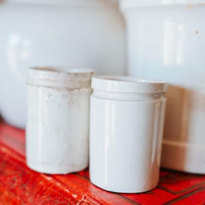 May include: Two small, white ceramic jars with lids sit on a vibrant red surface, with larger white ceramic containers in the background. The jars have a cylindrical shape and a slightly aged appearance, suggesting a vintage aesthetic.