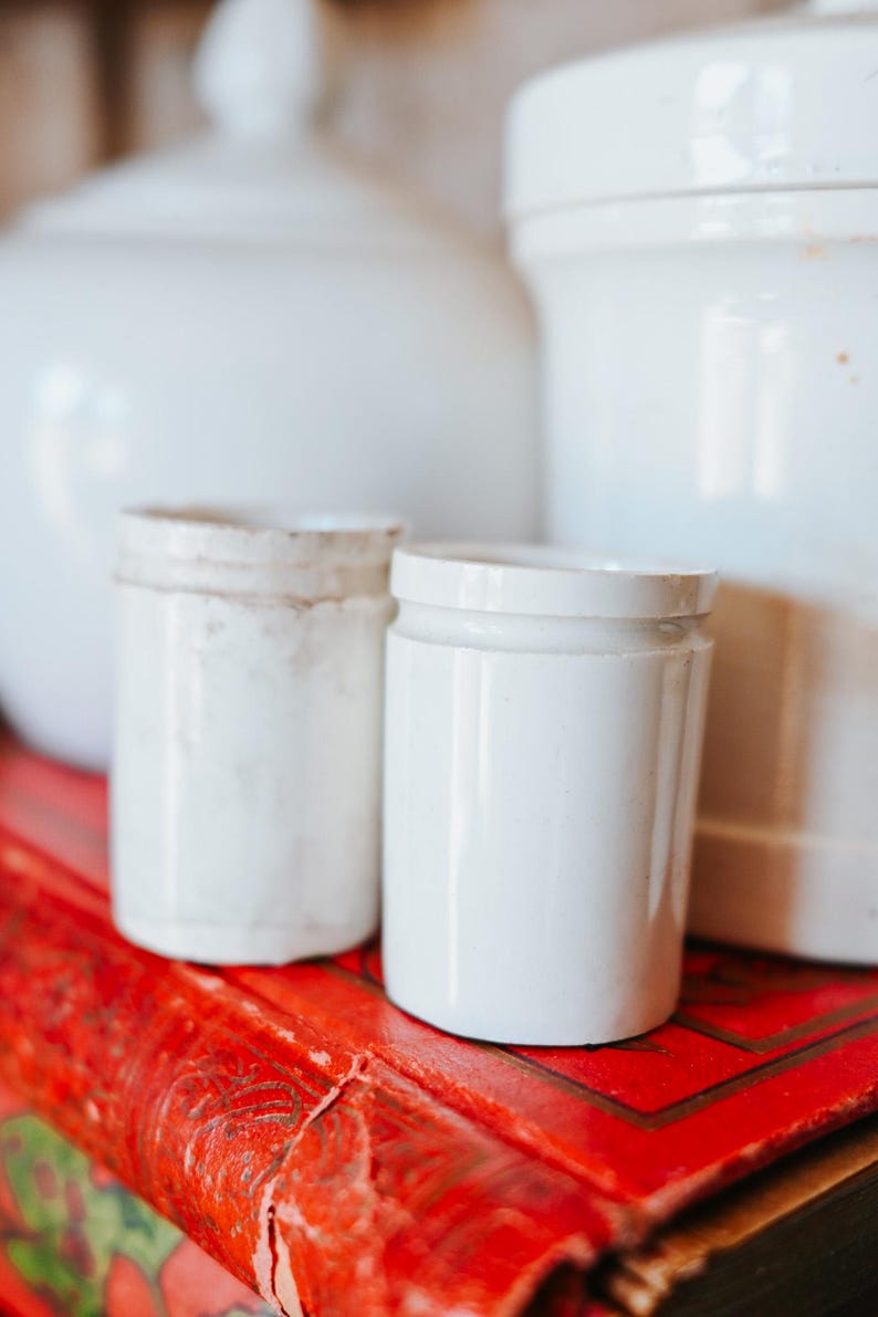 May include: Two small, white ceramic jars with lids sit on a vibrant red surface, with larger white ceramic containers in the background. The jars have a cylindrical shape and a slightly aged appearance, suggesting a vintage aesthetic.