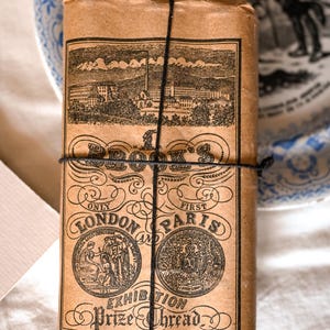 May include: A rectangular package wrapped in brown paper, tied with black string. The label features black ink illustrations of a factory, two circular medallions, and text reading BROOK'S, LONDON AND PARIS EXHIBITION, and Prize Thread.
