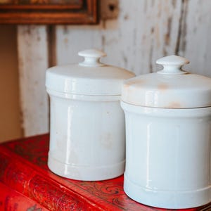 May include: Two white ceramic jars with lids, sitting on a stack of red books with floral designs. The jars have a classic shape and a small knob on top of the lid. The background includes a framed picture and a white wooden wall.