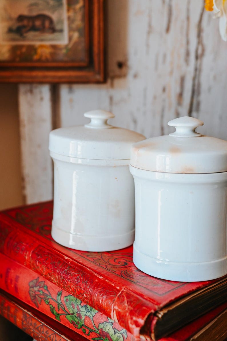 May include: Two white ceramic jars with lids, sitting on a stack of red books with floral designs. The jars have a classic shape and a small knob on top of the lid. The background includes a framed picture and a white wooden wall.