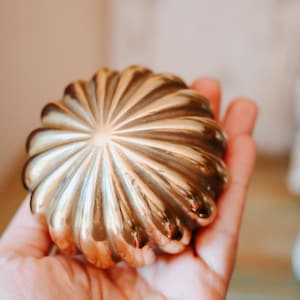 Pode incluir: Um objeto redondo de metal dourado com um design radial canelado, apresentando numerosas cristas que convergem no centro. A superfície tem um acabamento polido e reflexivo com variações subtis de tom ao longo do exterior curvo.