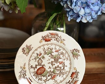 Puede incluir: Un plato redondo de cerámica con base color crema que presenta un patrón floral y arquitectónico en tonos marrón, naranja y verde. El plato tiene un borde festoneado y se exhibe sobre un soporte negro, mostrando intrincados motivos botánicos.