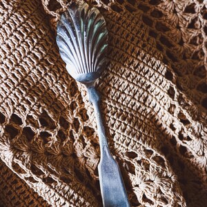 Könnte beinhalten: Ein silberner Servierlöffel mit einer muschelförmigen Schale und einem langen, flachen Griff. Der Löffel liegt auf einer beigefarbenen, gehäkelten Oberfläche, was einen Kontrast in den Texturen erzeugt. Das Design des Löffels deutet auf einen Vintage- oder Antikstil hin.