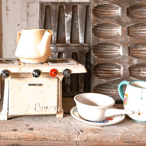 May include: A vintage-style kitchen scene featuring a cream-colored toy stove with black and red knobs, a teapot, a cup and saucer, and a floral-decorated mug. A metal madeleine pan is in the background.