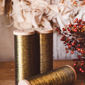 May include: Three spools of gold-colored thread with wooden ends are arranged on a wooden surface. One spool lies horizontally, while two stand upright. The background features a blurred fabric and a branch with red berries.