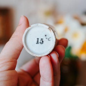 May include: A small, cylindrical, antique ceramic container with a white interior and a faded, aged exterior. The number "15" is printed in black on the bottom. The container is held in a person's hand, with a blurred floral background.