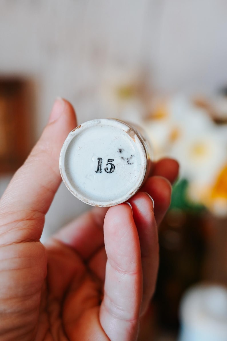 May include: A small, cylindrical, antique ceramic container with a white interior and a faded, aged exterior. The number "15" is printed in black on the bottom. The container is held in a person's hand, with a blurred floral background.